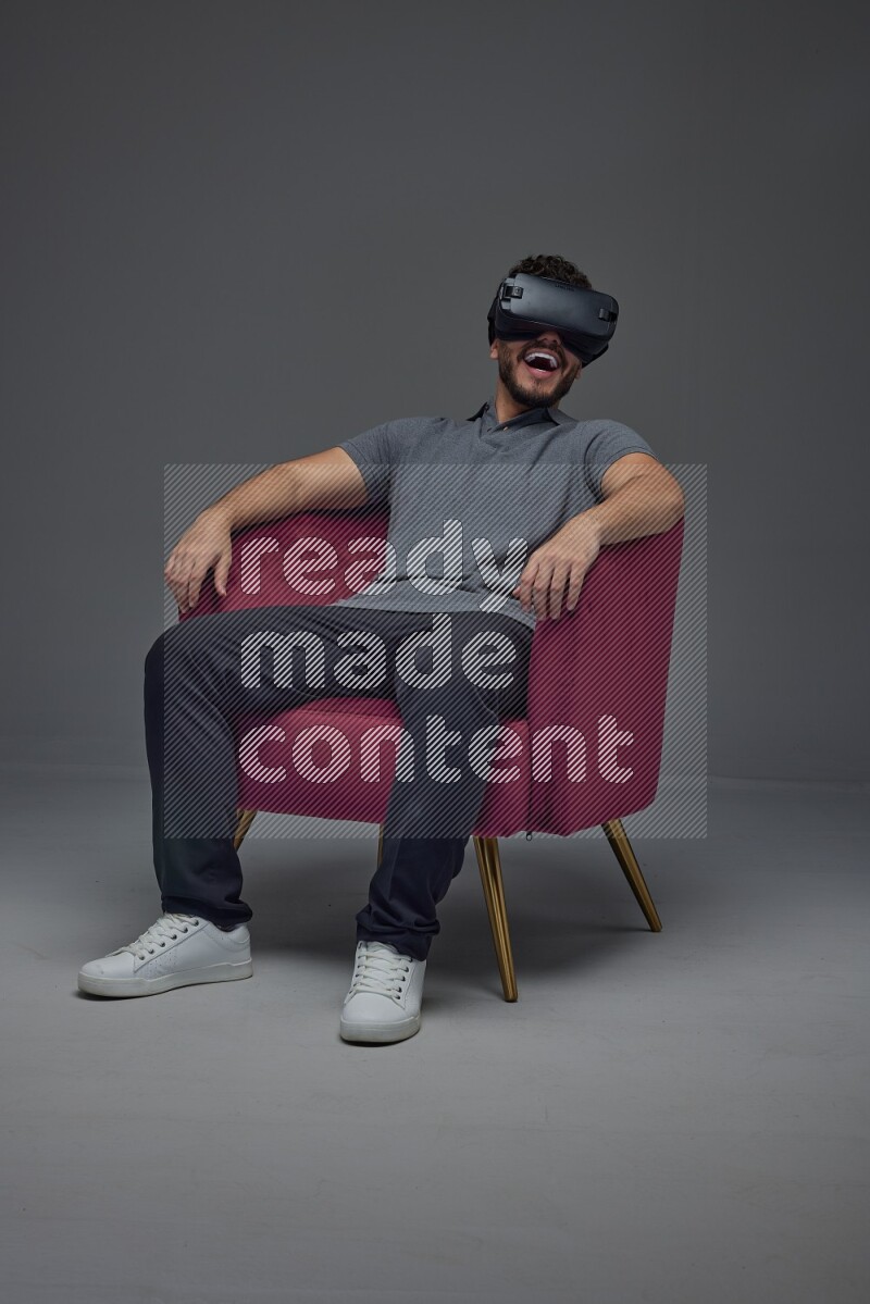 A man wearing casual and wearing VR while sitting on a burgundy chair eye level on a gray background