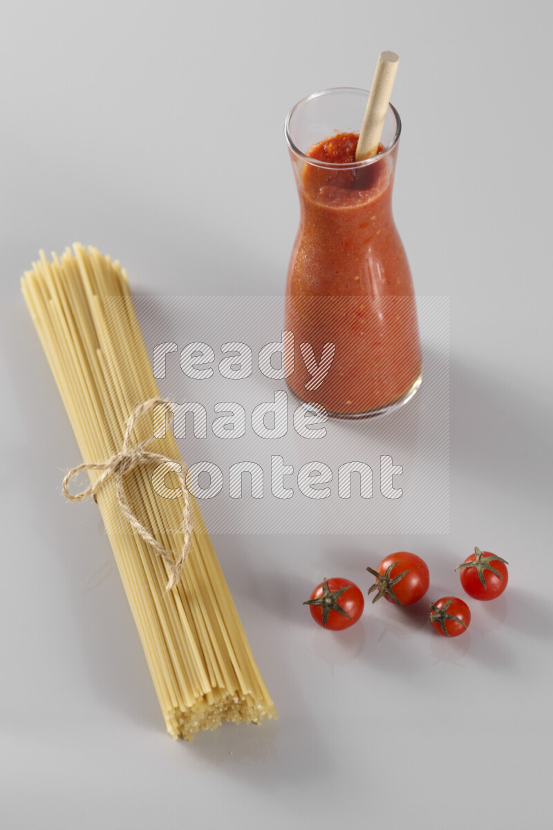 Raw pasta with tomatoe pasta with different ingredients such as cherry tomatoes, basil, garlic, bay laurel, cardamom, white pepper, black pepper, red chilis and wheat stalks on light grey background