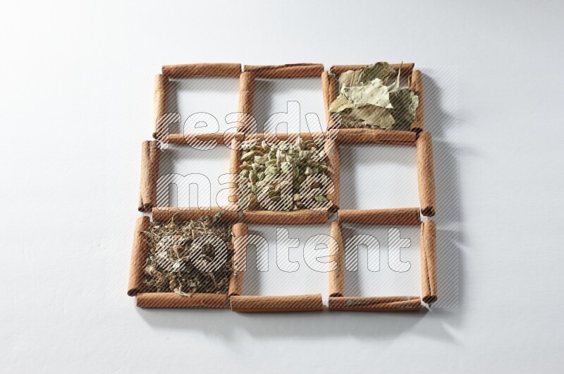 9 مربعات من أعواد القرفة ممتلئة بالهيل في المنتصف محاطة بجوزة الطيب والقرفة وأوراق الغار والقرنفل والكمون والريحان المجفف والزنجبيل المجفف واليانسون النجمي على أرضية بيضاء