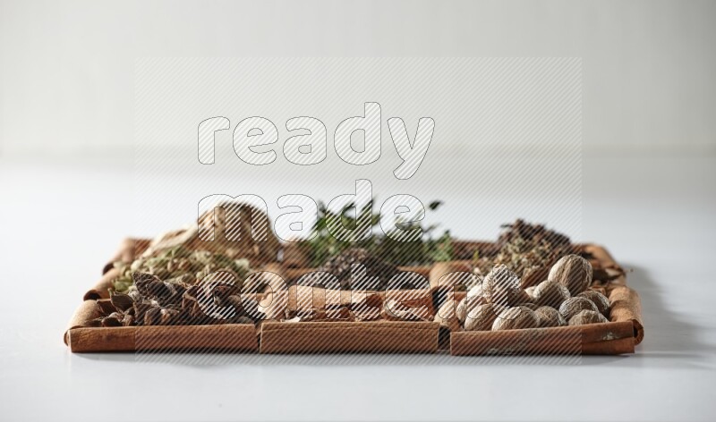9 squares of cinnamon sticks full of tea in the middle surrounded by nutmeg, dried mint, cloves, dried basil, dried ginger, cinnamon, star anise and cardamom on white flooring