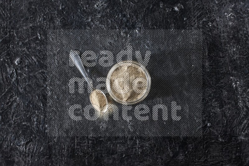 A glass jar and a metal spoon full of white pepper powder on textured black flooring