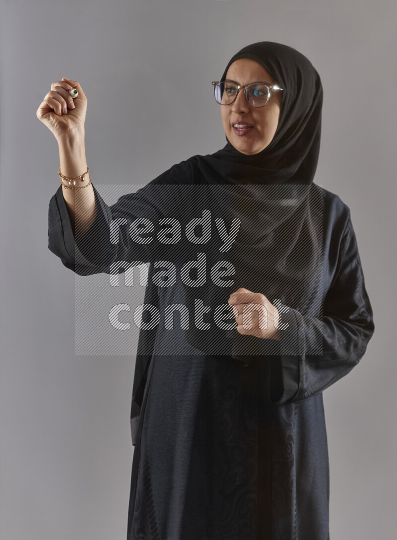 Woman in black abaya, hijab and glasses holding a marker pen to write on a transparent board with different reactions and expressions on grey background
