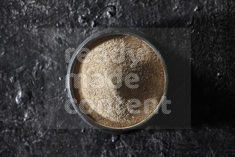 Black pottery bowl full of white pepper powder on textured black flooring