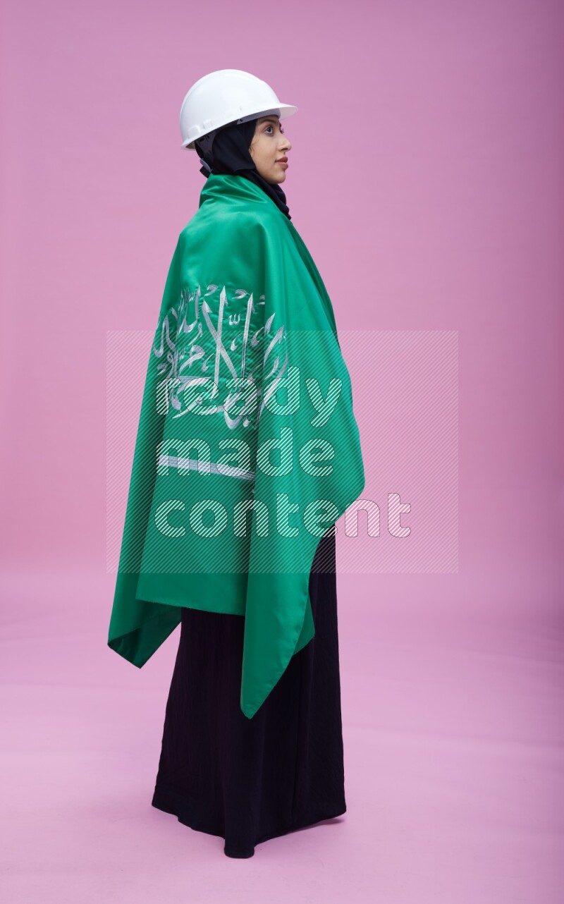 Saudi woman wearing Abaya with engineer vest and helmet standing holding Saudi flag on pink background