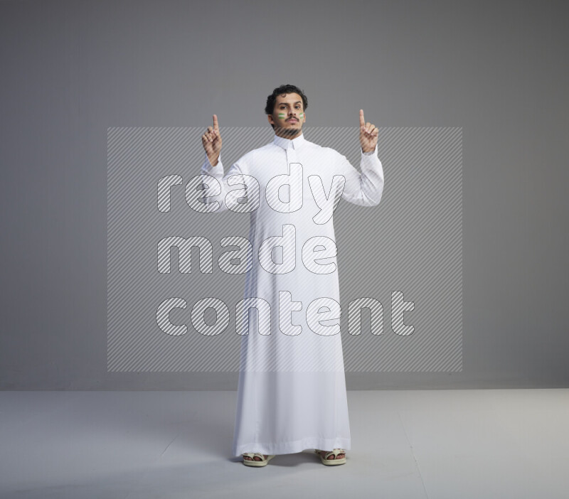 A Saudi man standing wearing thob with face painting interacting with the camera on gray background