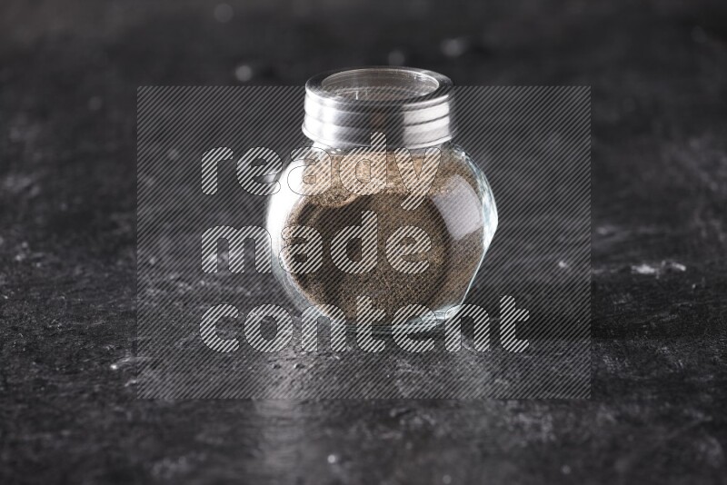 A glass spice jar full of black pepper powder on textured black flooring
