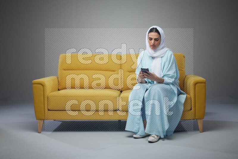 A Saudi woman wearing a light blue Abaya and white head scarf sitting on a yellow sofa and texting on her phone on a grey background