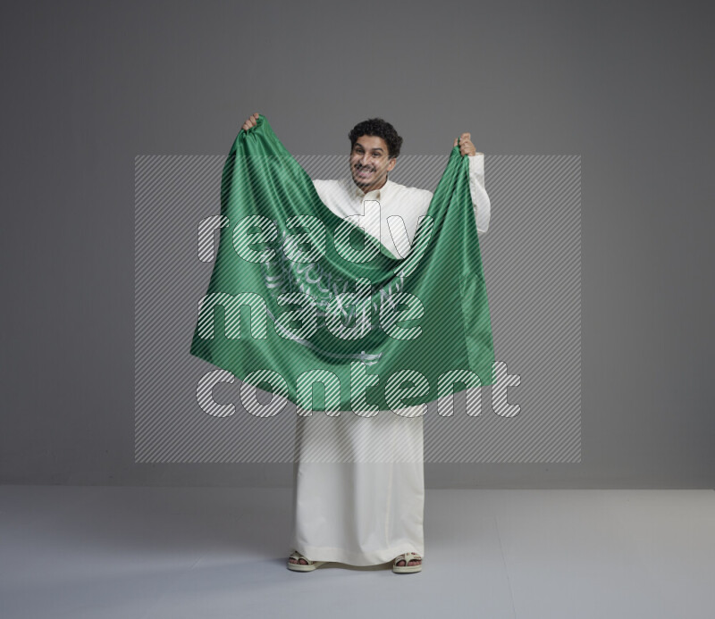 A Saudi man standing wearing thob holding big Saudi flag on gray background