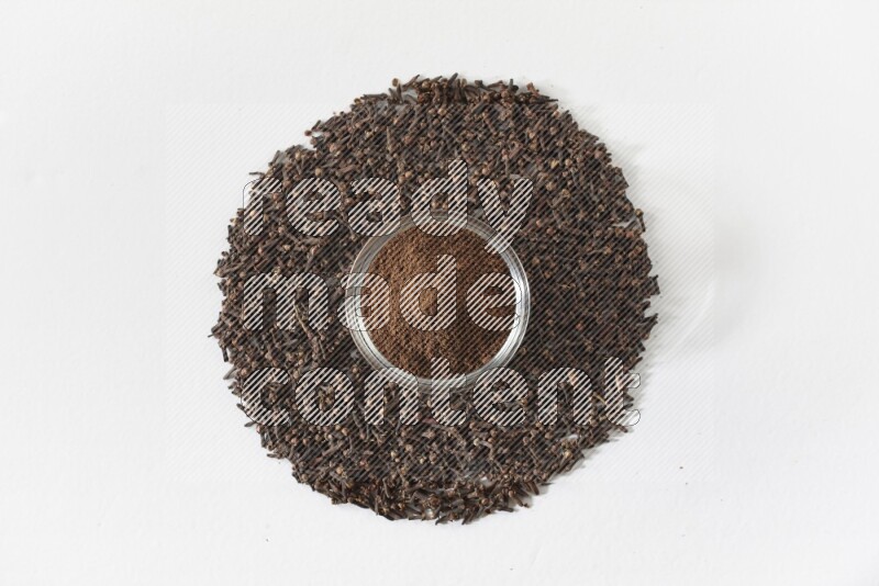 وعاء زجاجي ممتلئ ببودرة القرنفل وحبوب القرنفل متناثرة على أرضية بيضاء