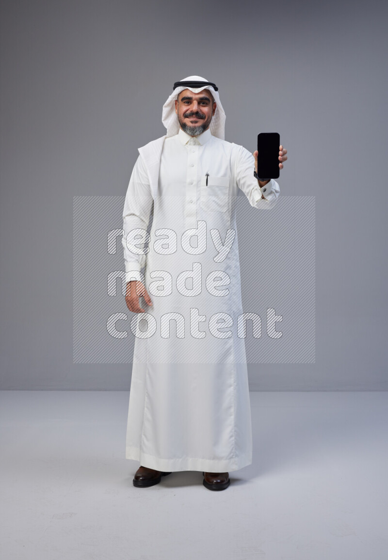 Saudi man Wearing Thob and white Shomag standing showing phone to camera on Gray background