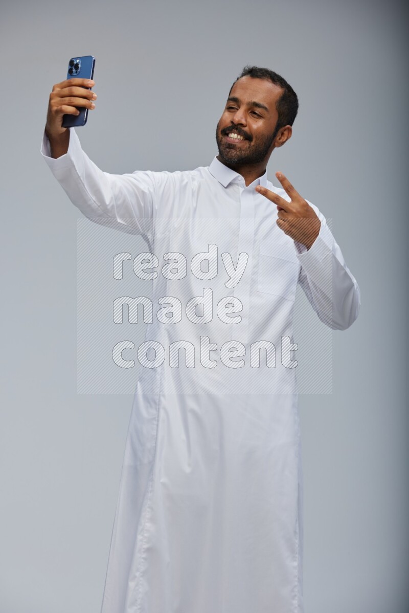 Saudi man wearing Thob standing taking selfie on Gray background