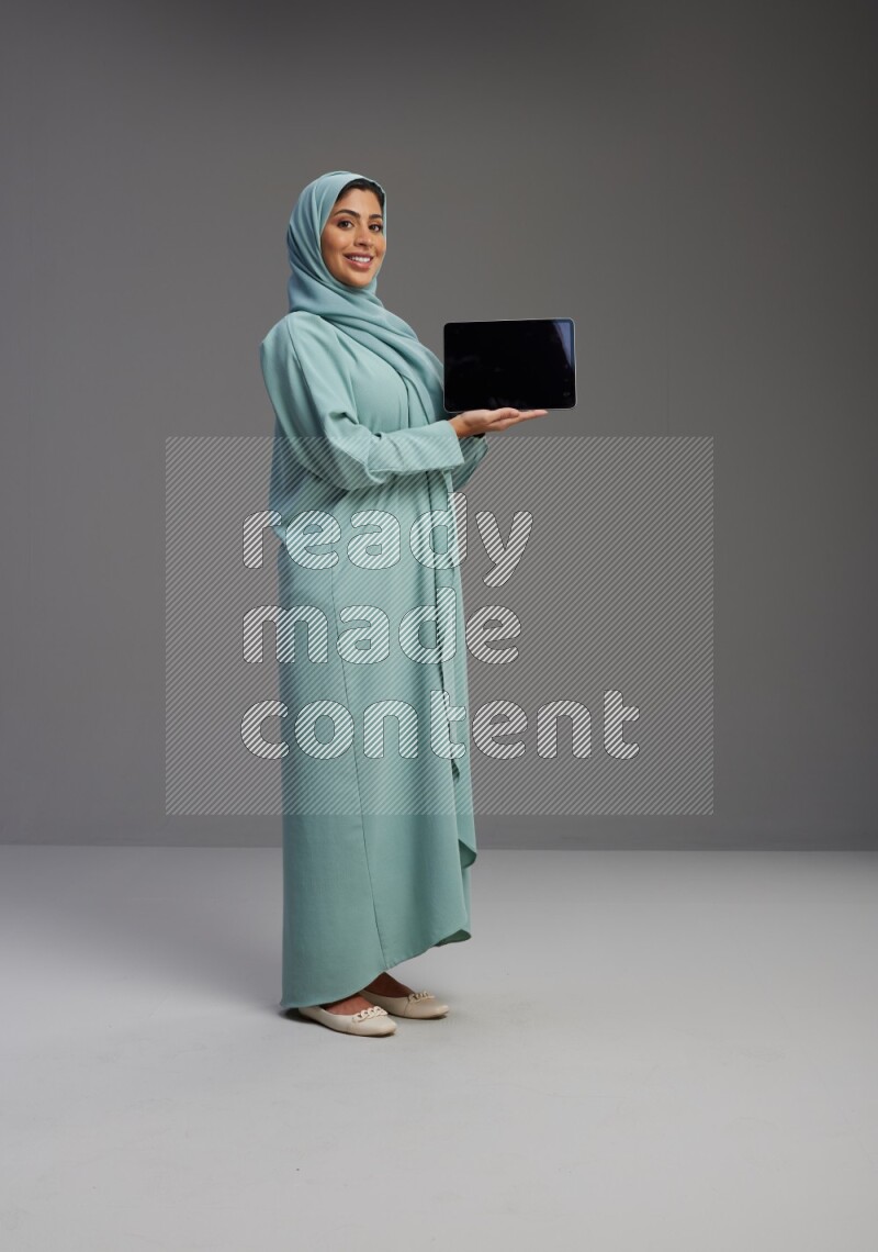 Saudi Woman wearing Abaya standing showing tablet to camera on Gray background