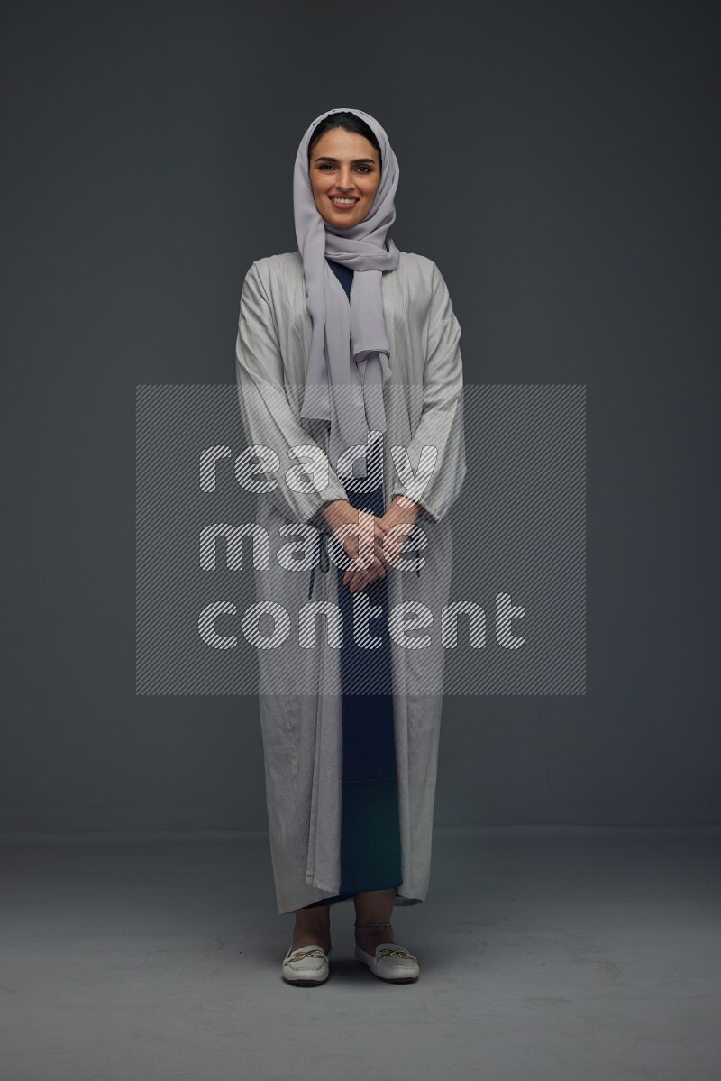A Saudi woman wearing a light gray Abaya and head scarf standing and making multi poses eye level on a grey background