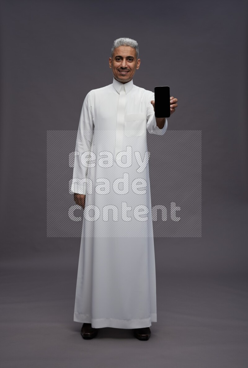 Saudi man wearing thob standing showing phone to camera on gray background