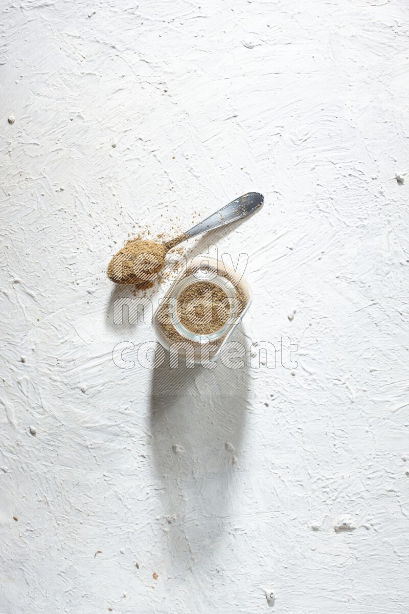 A glass spice jar and metal spoon full of cumin powder on textured white flooring
