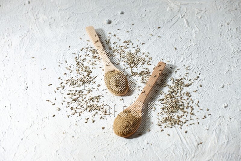 2 Wooden spoons full of cumin powder and cumin seeds on textured white flooring