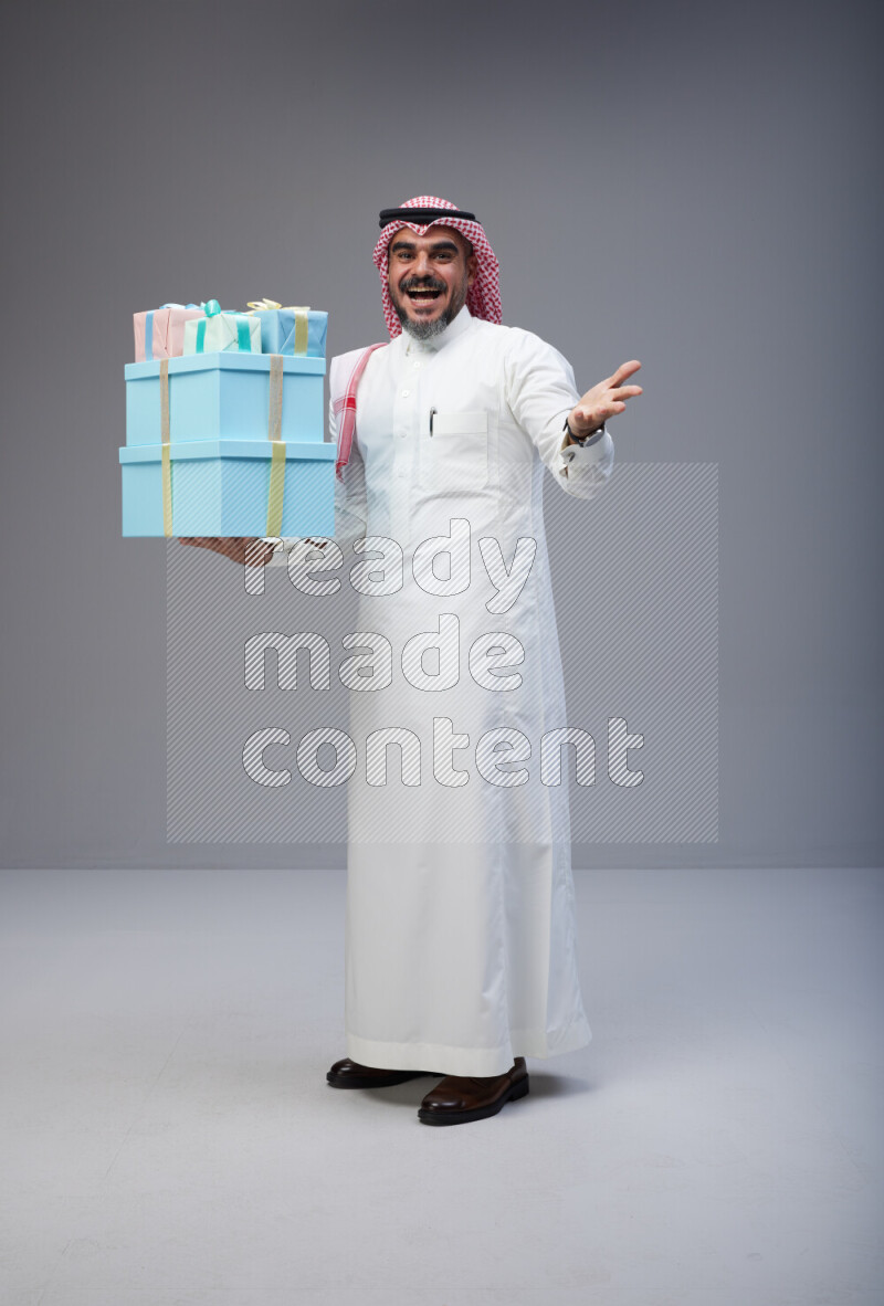 Saudi man Wearing Thob and red Shomag standing holding gift box on Gray background