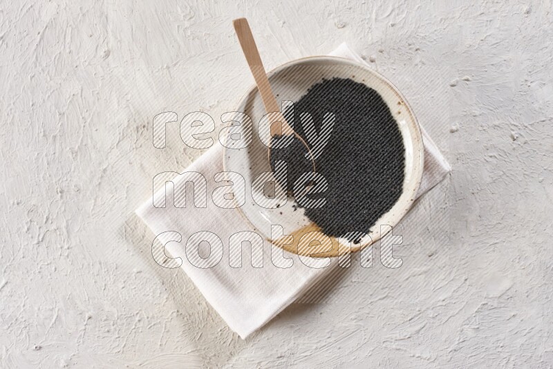 A multicolored pottery plate full of black seeds with a wooden spoon full of the seeds on a napkin on a textured white flooring