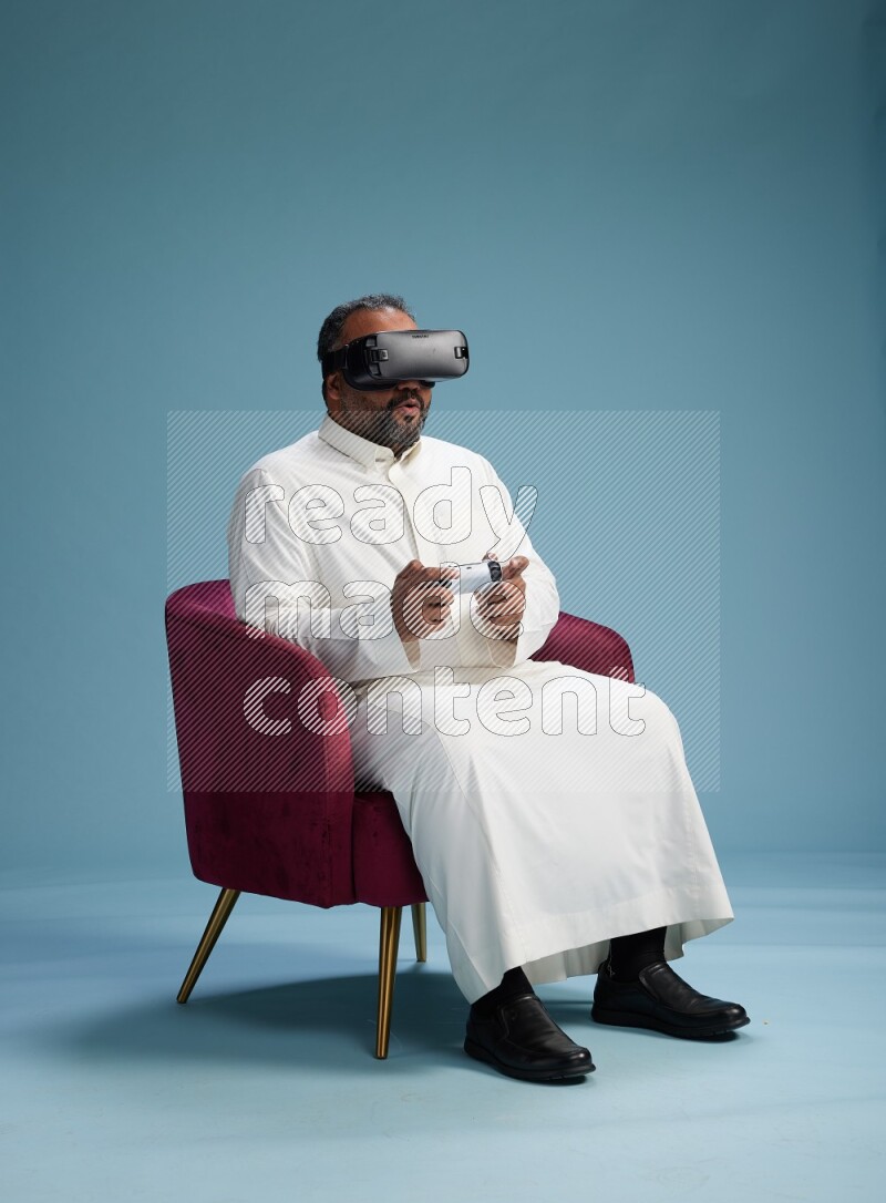Saudi Man without shimag sitting on chair  gaming with VR and joystick on blue background