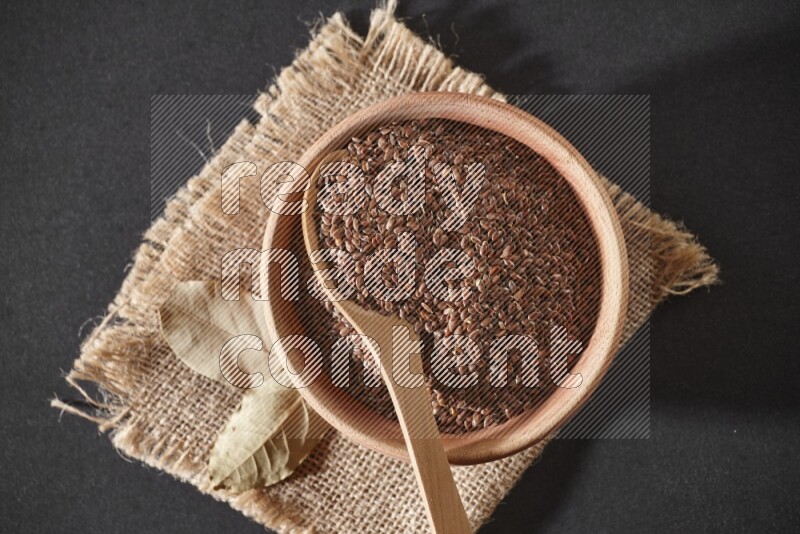 A wooden bowl full of flaxseeds with wooden spoon full of the seeds on it on burlap fabric on a black flooring
