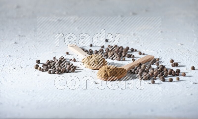 Wooden spoons full of allspice powder and allspice whole balls spreaded on a textured white flooring