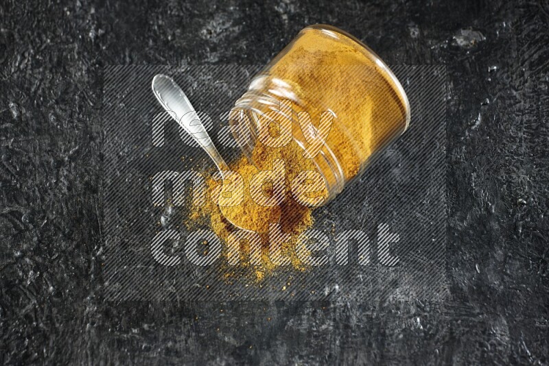 A flipped glass jar and a metal spoon full of turmeric powder and powder spilled out of it on textured black flooring