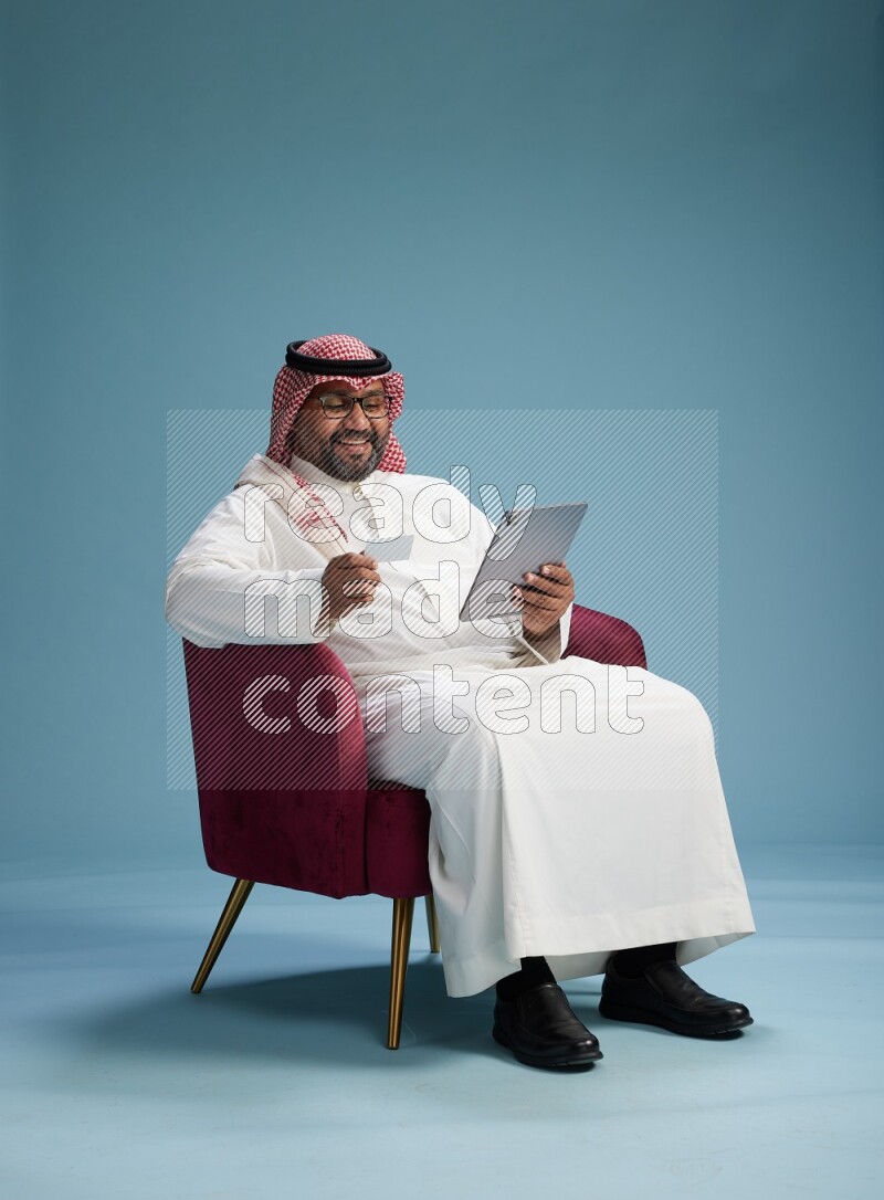 Saudi Man with shimag sitting on chair holding ATM card while working on tablet on blue background
