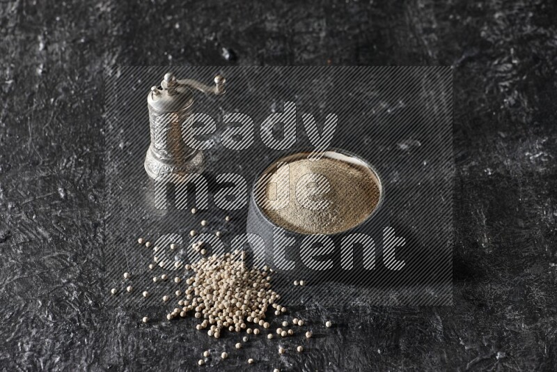 Black pottery bowl full of white pepper powder with pepper beads and metal pepper grinder on textured black flooring