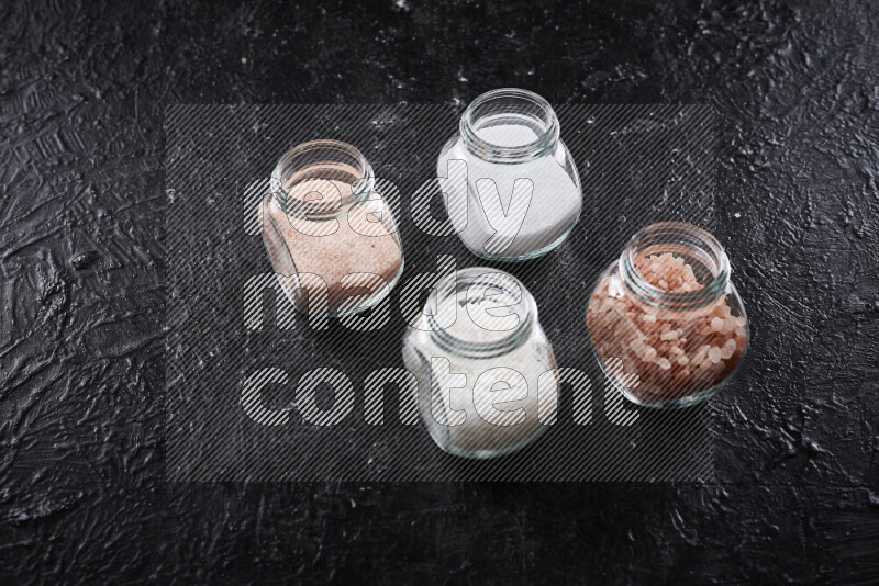 4 glass jars filled with table salt, coarse sea salt, fine himalayan salt and coarse himalayan salt on black background