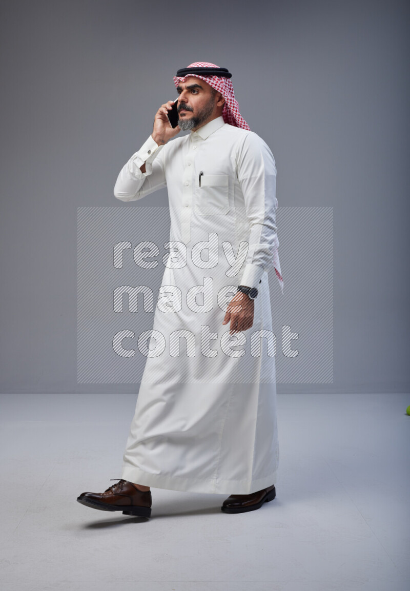 Saudi man Wearing Thob and red Shomag standing talking on phone on Gray background