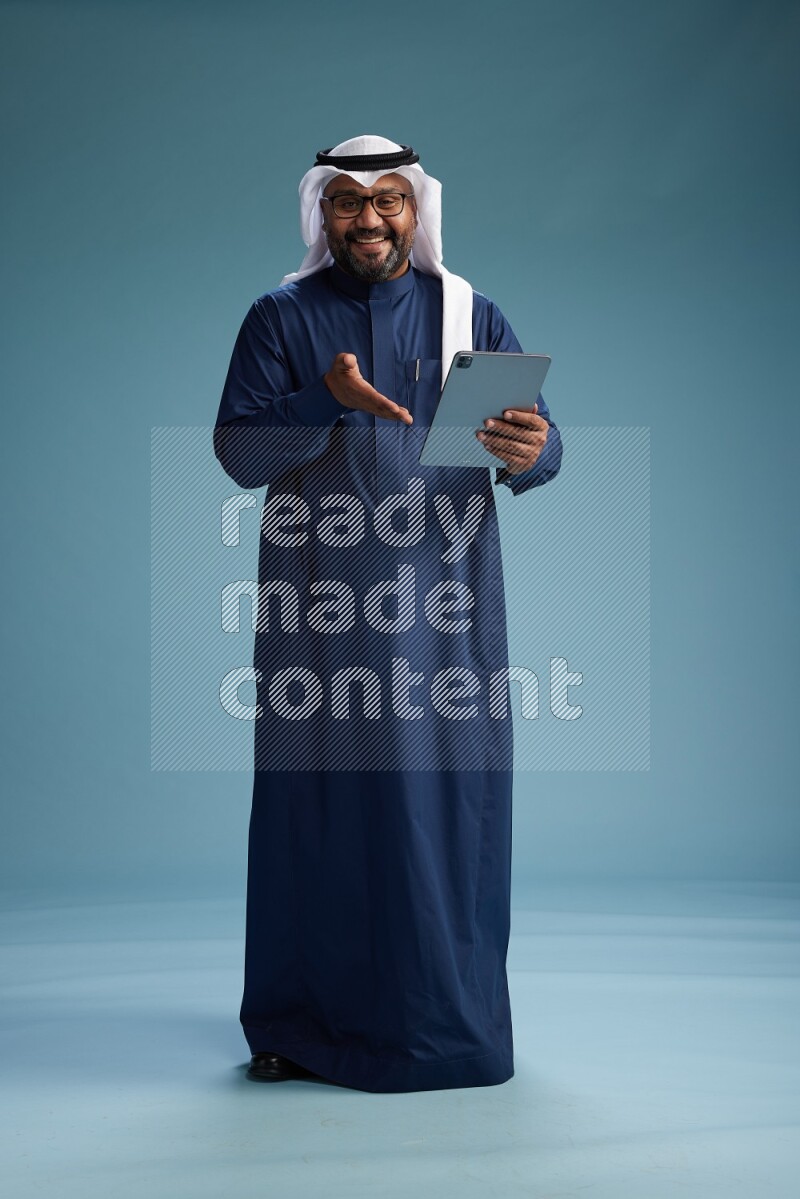 Saudi Man with shimag Standing holding ATM card while working on tablet on blue background
