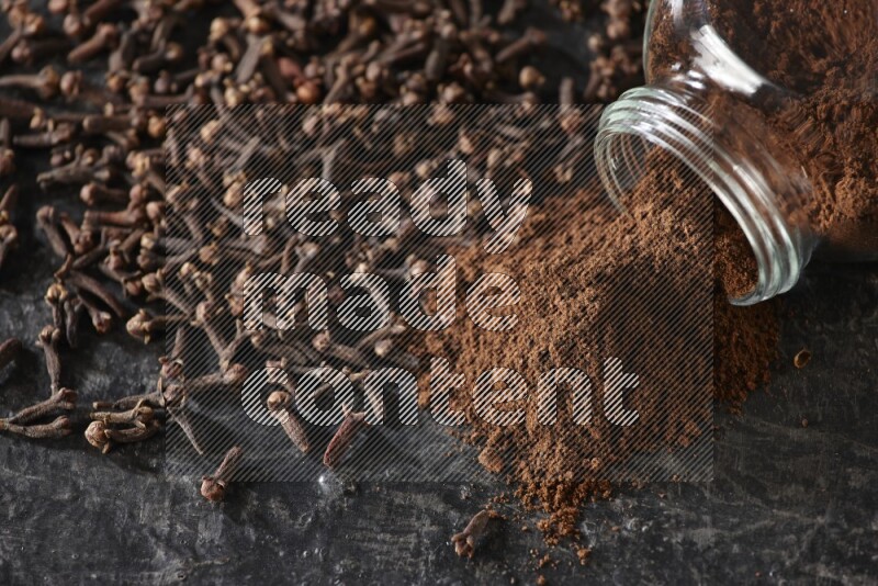 A flipped glass spice jar full of cloves powder and powder came out of it with cloves spread on textured black flooring