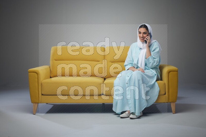 A Saudi woman wearing a light blue Abaya and a white head scarf sitting on a yellow sofa and talking in the phone while making different poses eye level on a grey background