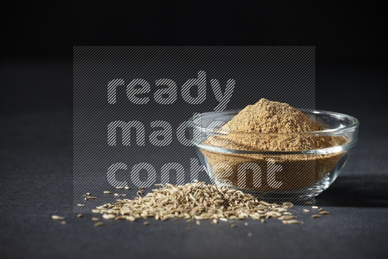 A glass bowl full of cumin powder with cumin seeds beside it on black flooring