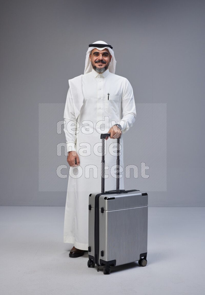 Saudi man wearing Thob and white Shomag standing holding Travel bag on Gray background
