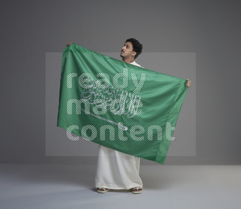 A Saudi man standing wearing thob holding big Saudi flag on gray background