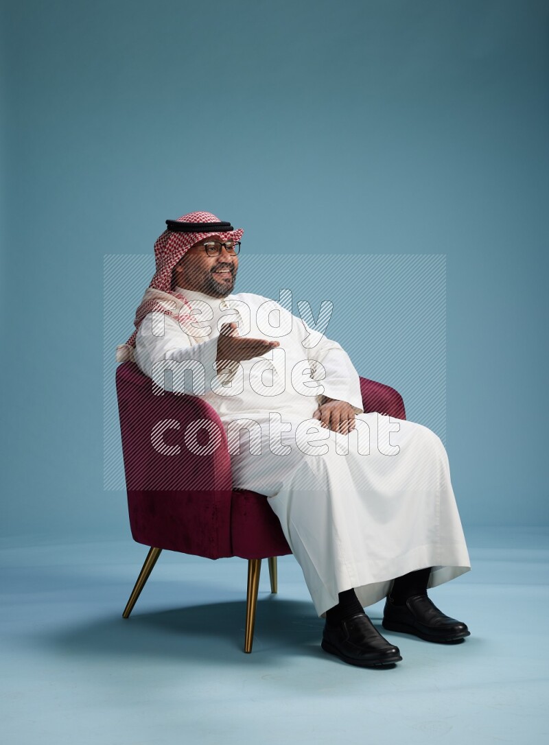 Saudi Man with shimag sitting on chair Interacting with the camera on blue background