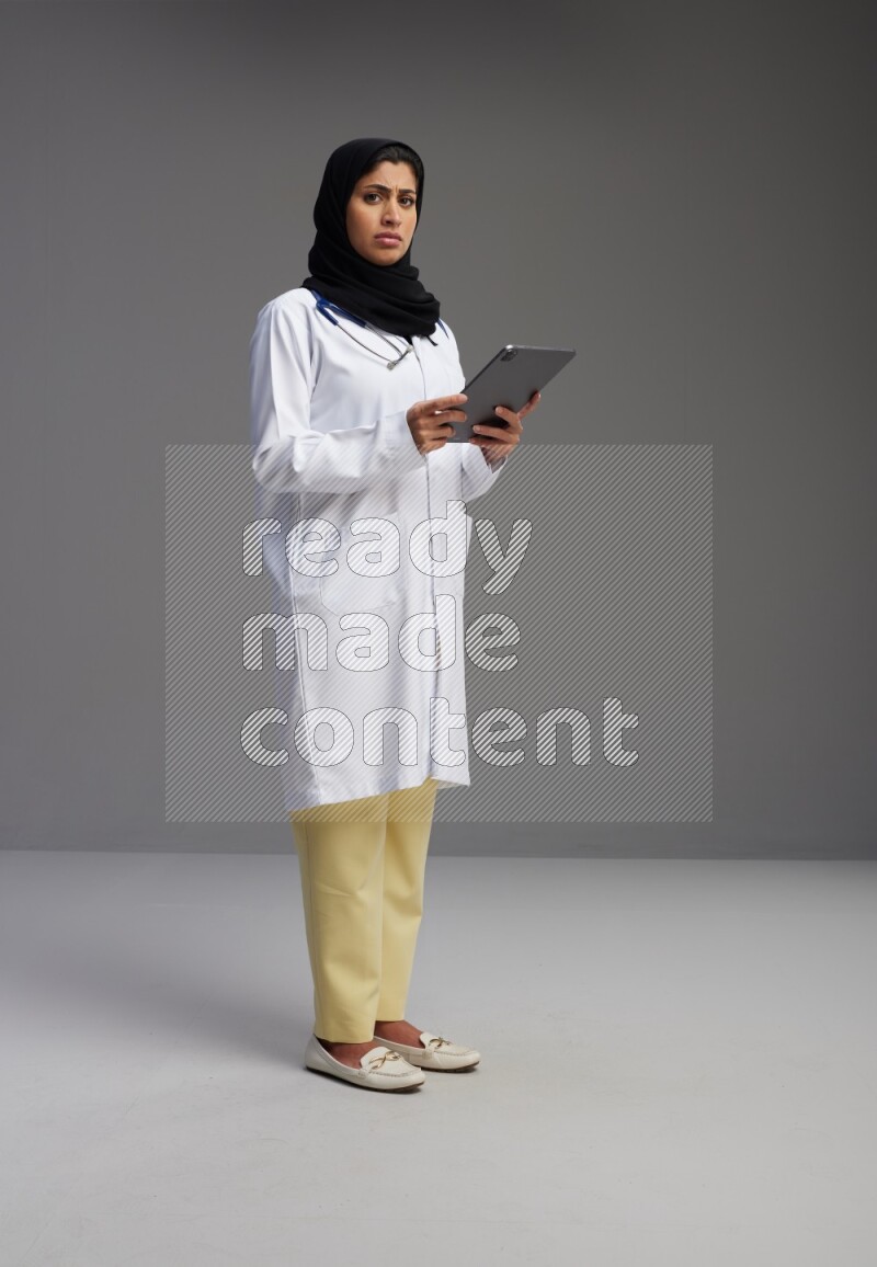 Saudi woman wearing lab coat with stethoscope standing working on tablet on Gray background