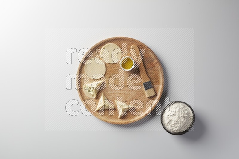 two closed sambosas and one open sambosa filled with cheese while flour, and oil with oil brush aside in a wooden dish on a white background