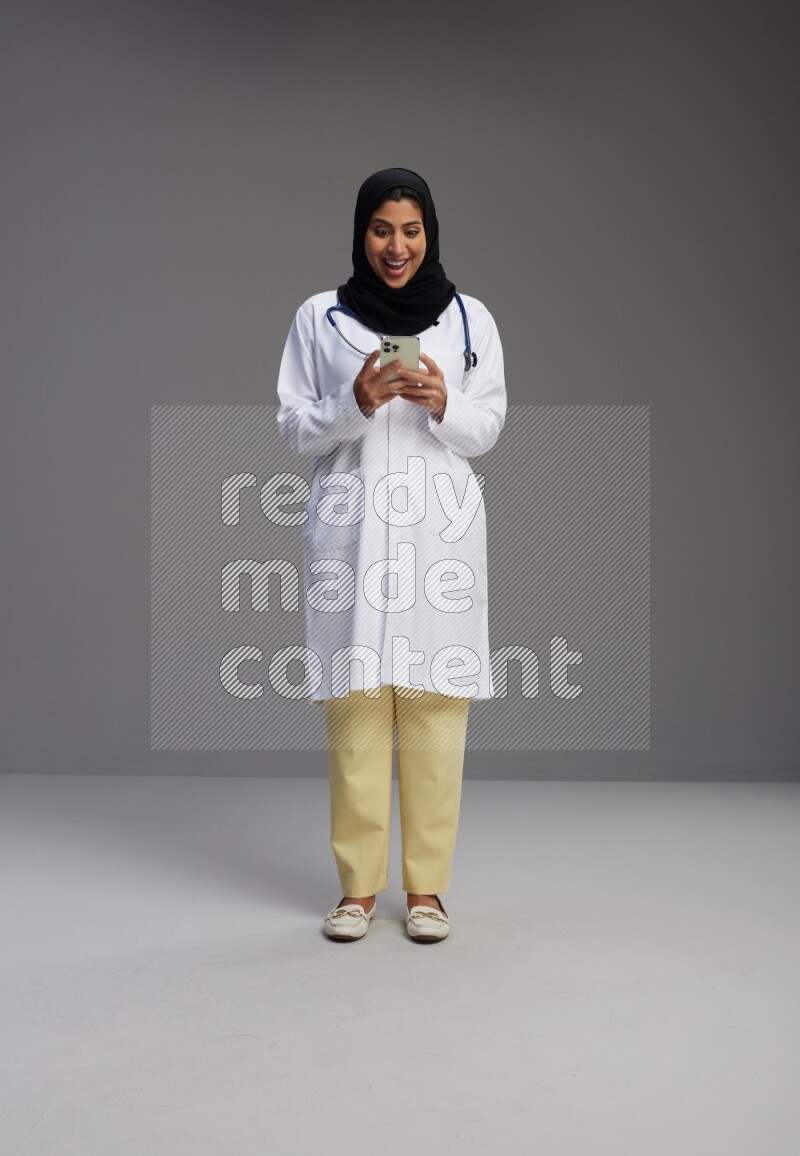 Saudi woman wearing lab coat with stethoscope standing texting on phone on Gray background