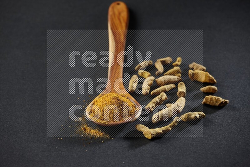 A wooden ladle full of turmeric powder with dried turmeric fingers on black flooring