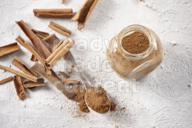 Herbal glass jar full cinnamon powder and a metal spoon surrounded by cinnamon sticks on a white background
