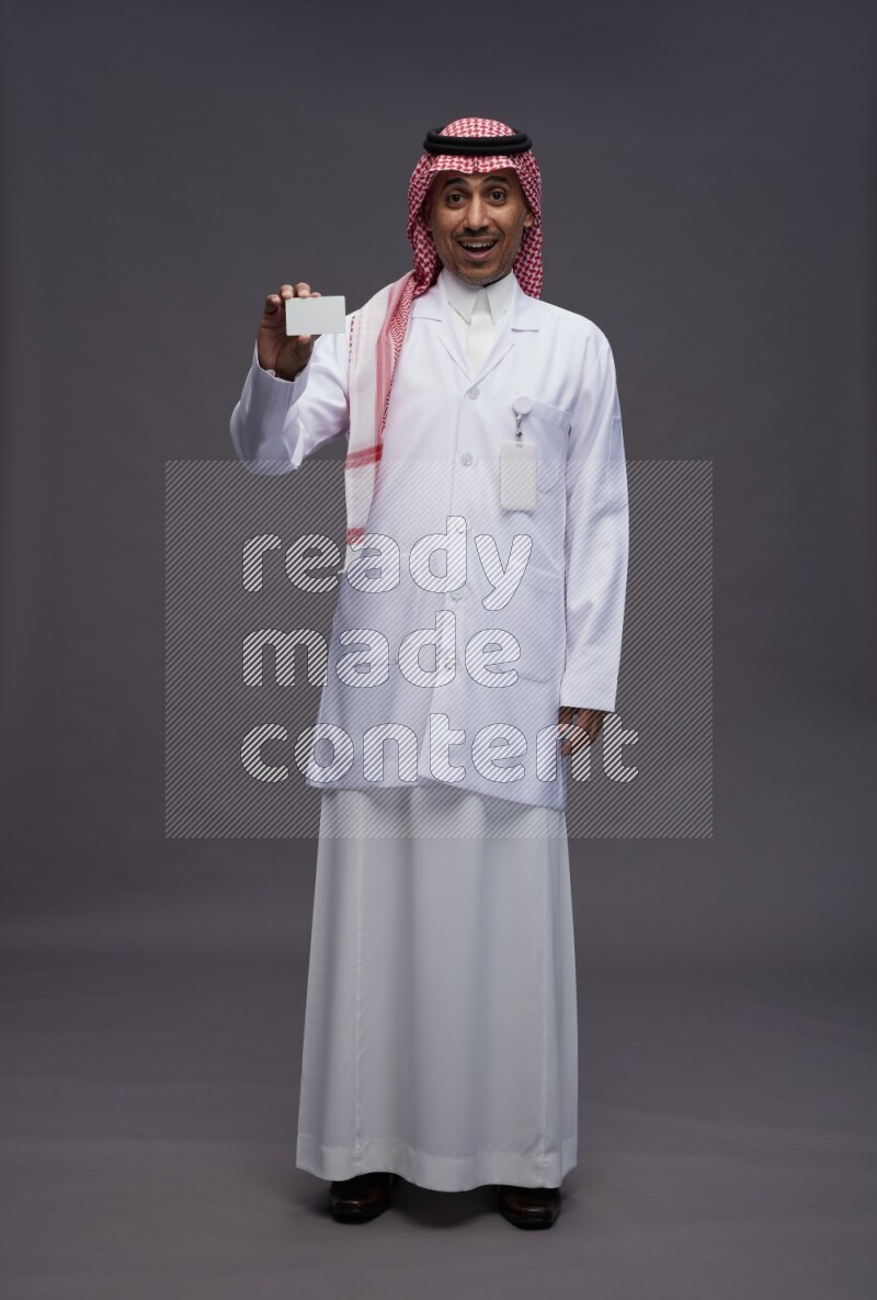 Saudi man wearing thob with lab coat and shomag with pocket employee badge standing holding ATM card on gray background