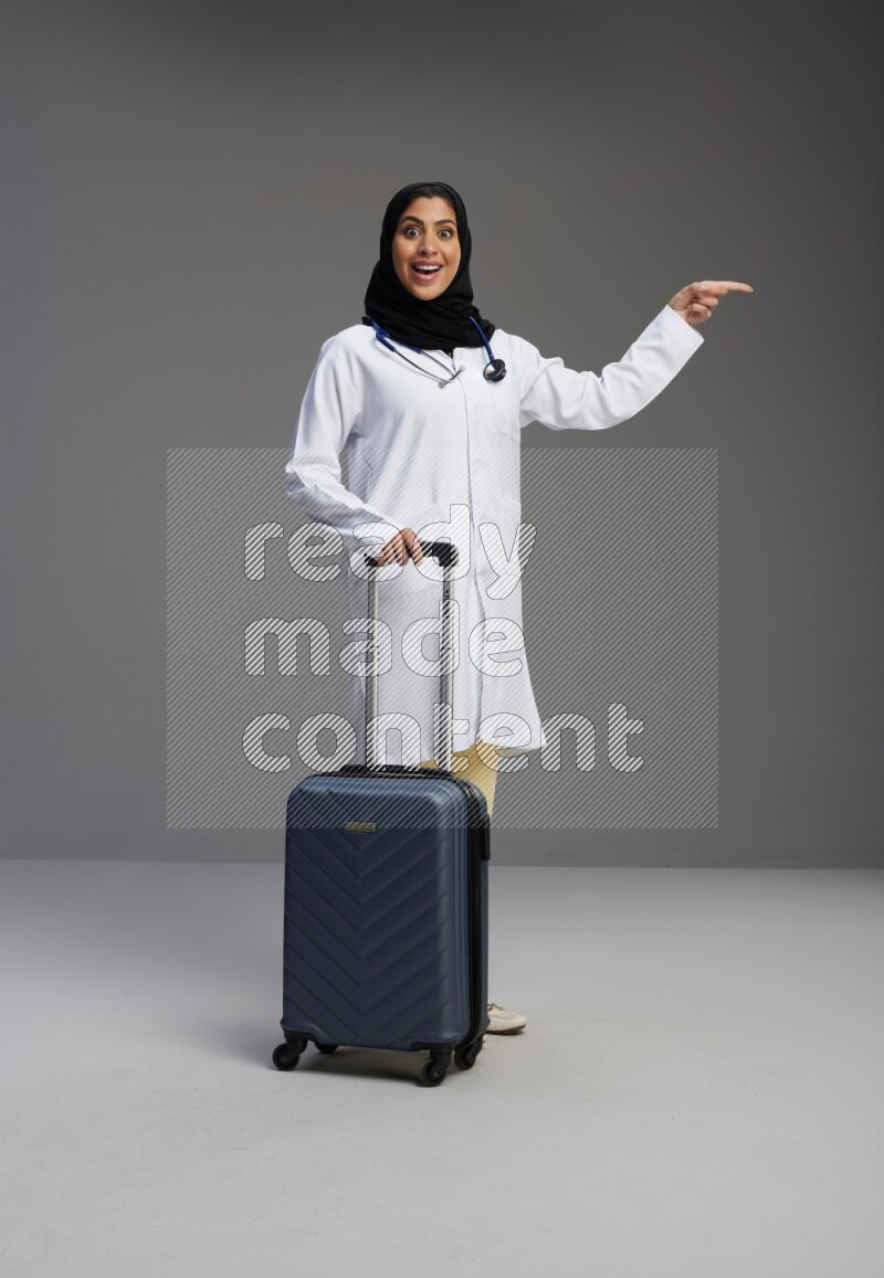 Saudi woman wearing lab coat with stethoscope standing holding Travel bag on Gray background