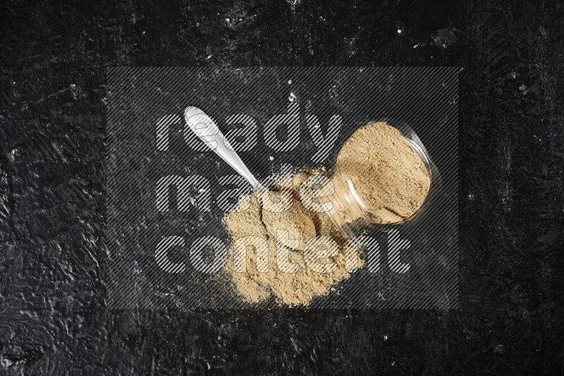 A glass jar full of ground ginger powder flipped with some spilling powder on black background