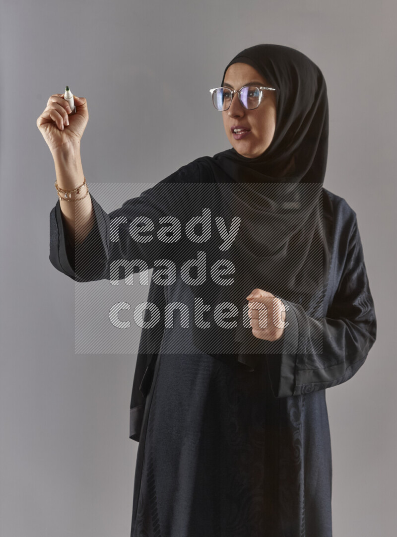 Woman in black abaya, hijab and glasses holding a marker pen to write on a transparent board with different reactions and expressions on grey background