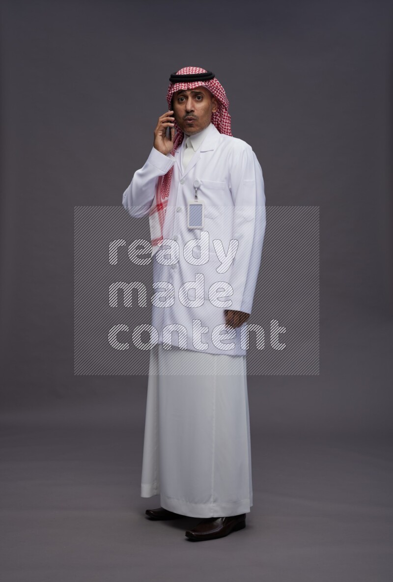 Saudi man wearing thob with lab coat and shomag with pocket employee badge standing talking on phone on gray background