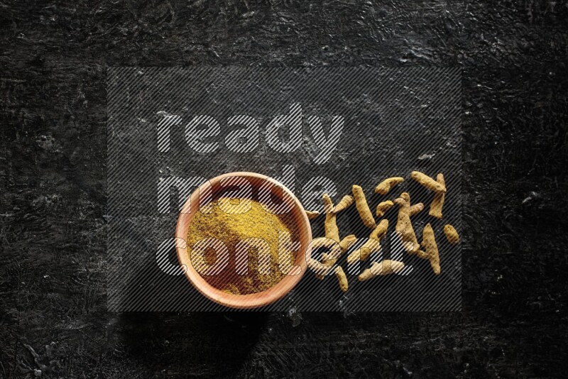 طبق خشبي ممتلئ ببودرة الكركم مع أصابع الكركم المجففة على خلفية سوداء