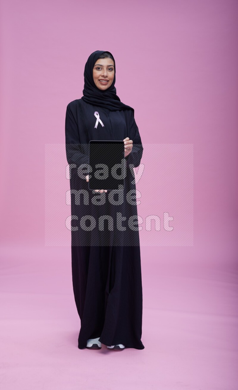 Saudi woman wearing pink ribbon on Abaya standing showing a tablet to camera on pink background