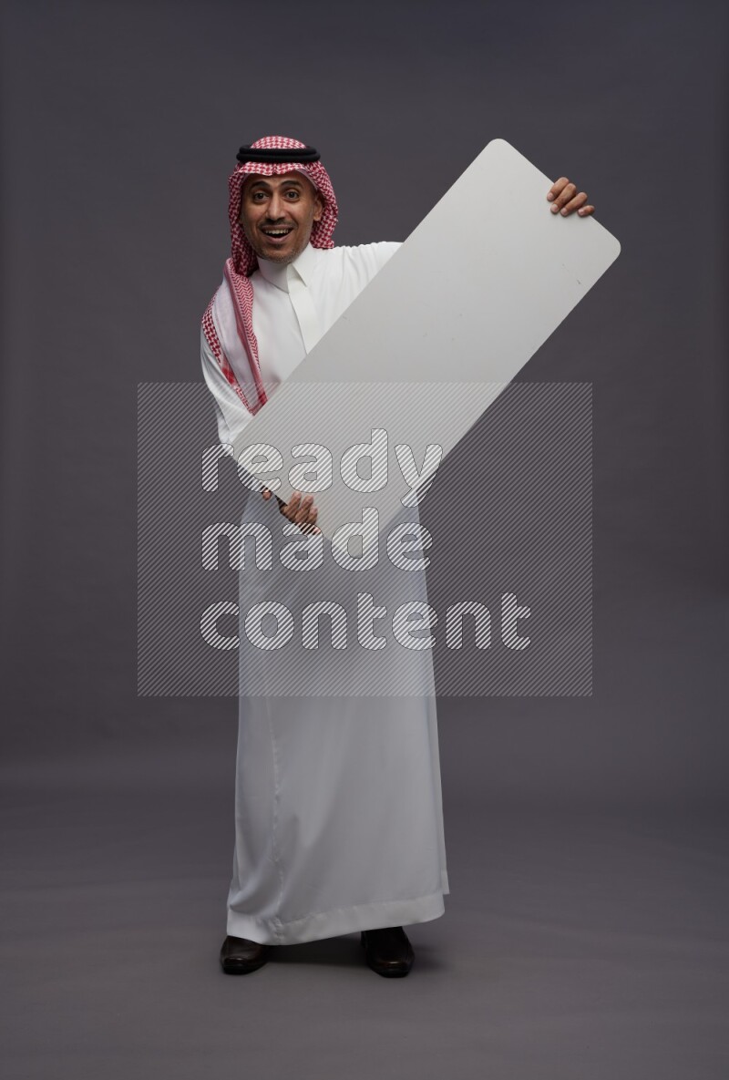 Saudi man wearing thob and shomag standing holding board on gray background